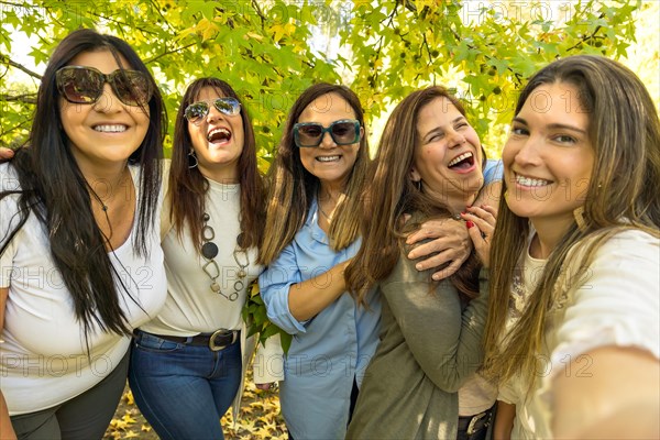 A group of five lively women are taking a selfie on a sunny day with a leafy tree in the background. Selfie with friends