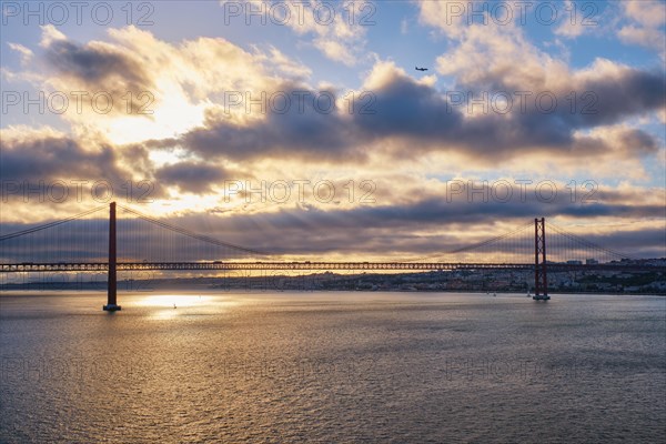 View of 25 de Abril Bridge famous tourist landmark of Lisbon connecting Lisboa and Almada on Setubal Peninsula over Tagus river on sunset with flying plane. Lisbon