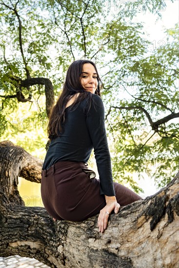 A woman sitting on a tree branch posing and looking at the camera in a public park