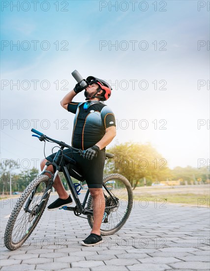 Thirsty cyclist on his bike drinking water outside. Tired cyclist drinking water outdoors