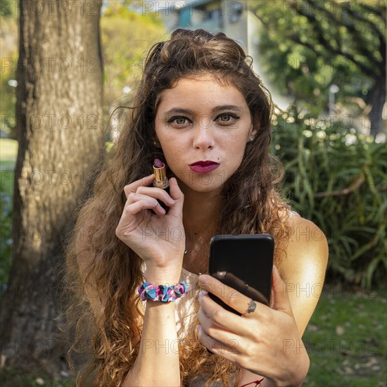 Beautiful young woman painting her lips using her smartphone to look her make up