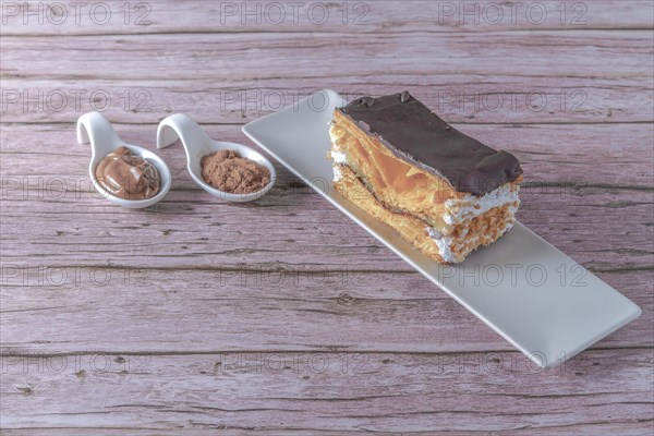Close-up of a mille-feuille filled with cream and almonds covered with chocolate on a white plate with ceramic spoons with chocolate and cocoa powder