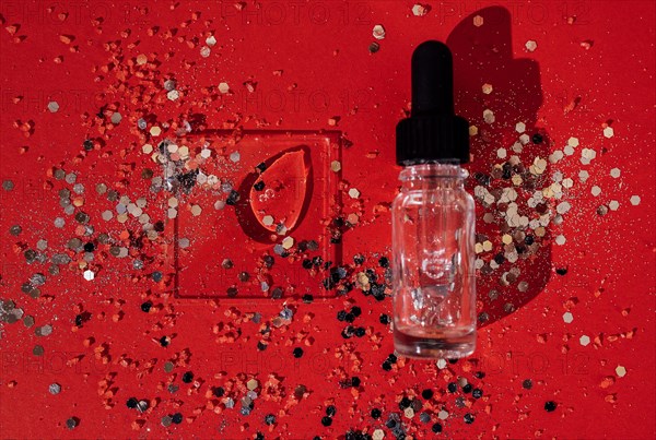 Transparent serum or oil glass bottle with pipette. Bright red background with silver glitters and confetti. Mock up. Concept of cosmetic and celebration. Copy space