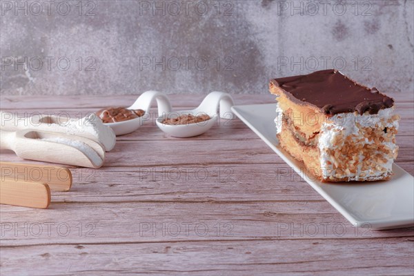 Close-up of a mille-feuille filled with cream and almonds covered with chocolate on a white plate with ceramic spoons with chocolate and cocoa powder