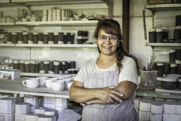 Adult woman potter with glasses and work apron smiling and looking at camera in her studio