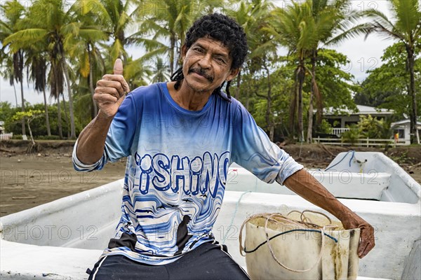 Man looking at camera with thumb up on his boat and fishing net. Background of palm trees out of focus