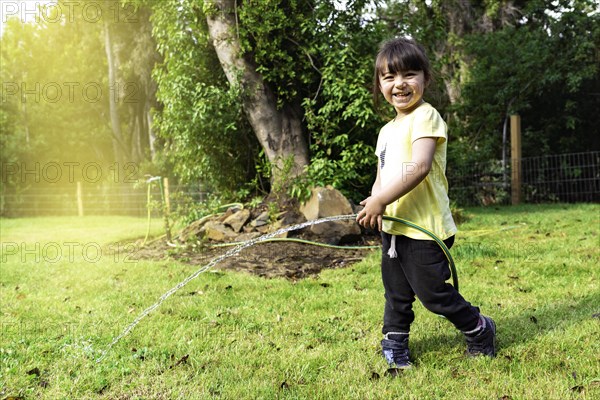 Portrait of happy smiling little child girl playing and watering garden grass with rubber strap and sunny summer in garden at home