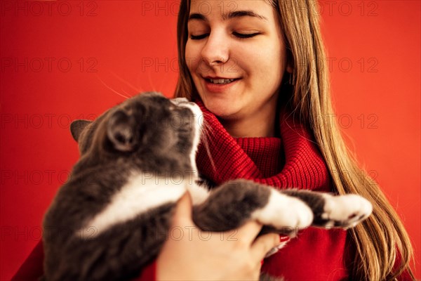 Portrait of blondy teenage girl in a red sweater holds a cat on her hand. Beautiful smiling girl with her pet. Close up. Copy space. Isolated on a red background