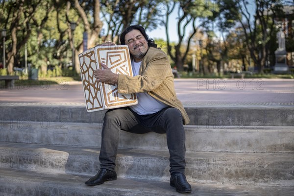Hispanic musician sitting in a park hugging his flamenco box