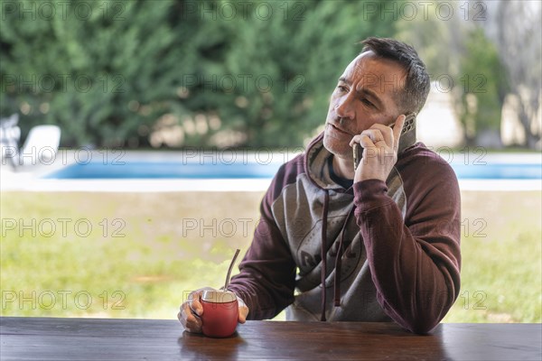 Serious adult man talking on the cellphone while drinking mate infusion at home