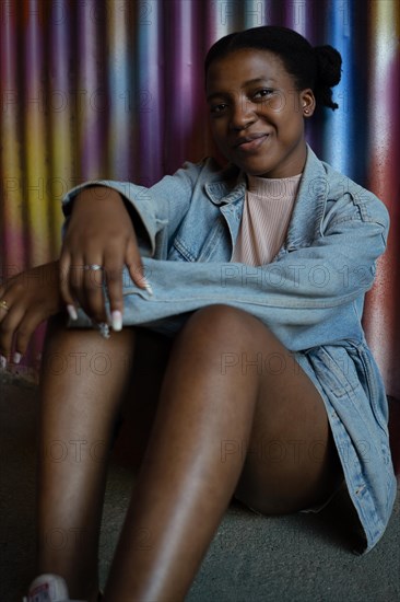 Pretty young African American girl smiling looking at the camera seated leaning her back against a colorful metal wall