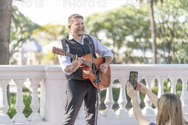 Street musician performing at a park and a girl from the audience taking pictures and filming him