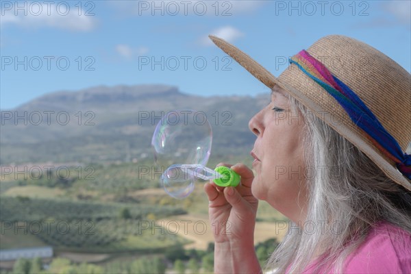 Older white-haired woman with hat seen in profile blowing soap bubbles with a mountain landscape in background