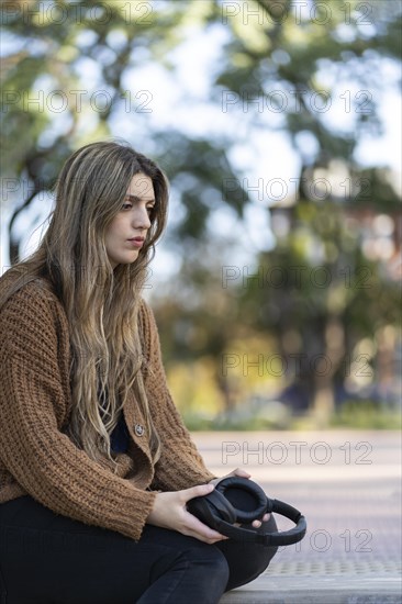 Young woman sitting in a park with a worried expression and a lost look while holding headphones