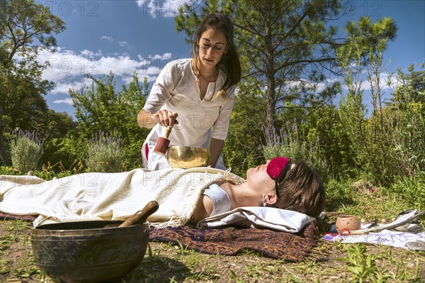 Woman therapist practicing sound healing therapy on a woman lying in a natural environment