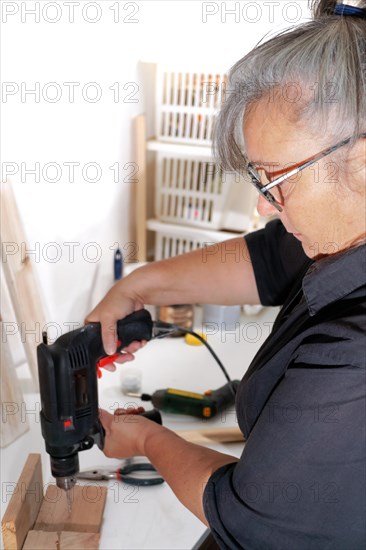 Woman see in profile drilling a wooden board with an electric drill on a white board in her workshop