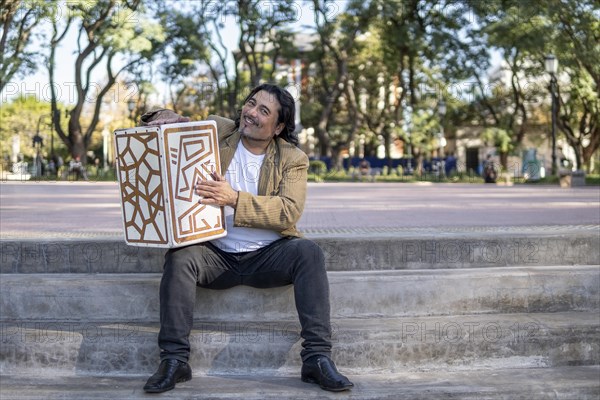 Hispanic musician showing in a park hugging his flamenco box