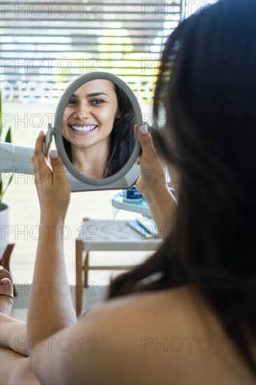 Young woman patient is looking in the mirror and admires her new smile after dental treatment in the dentistry clinic