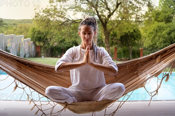 Blonde yogi woman with dreadlocks in prayer position sitting in a woven hammock