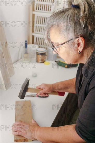 Woman seen in profile hammering a nail with a hammer into a wooden plank in her workshop