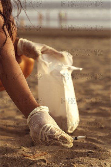 Volunteer girl collecting cigarette butts on the sand. Out-of-focus sunset background. Marine pollution destroying the environment