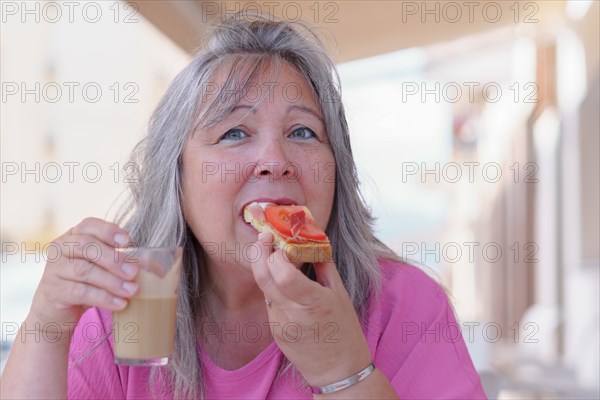 Close-up of an older white-haired woman drinking coffee with milk and eating a ham and tomato toast