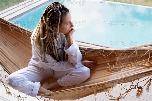 Blonde woman with dreadlocks thinking sitting in a woven hammock. Resting by the pool. Holidays. Relaxing moments