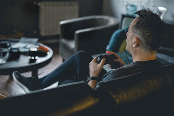 An adult man sitting on a sofa and holding a joystick while playing a video game on console