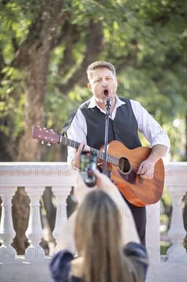 Street musician performing at a park and a girl from the audience taking pictures and filming him