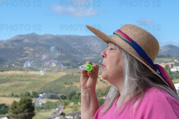 Older white-haired woman with hat seen in profile blowing soap bubbles with a mountain landscape in background