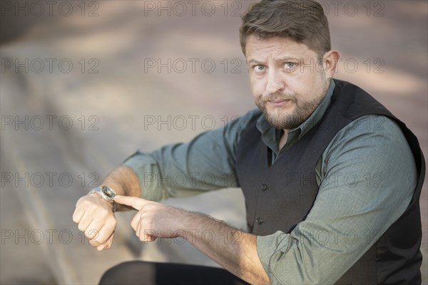 Man sitting in a park pointing at his watch looks disgusted at the camera. Delay concept