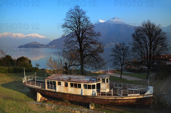 Old passenger ship on the field and trees with branches and alpine lake with snow-capped mountain in Lombardy
