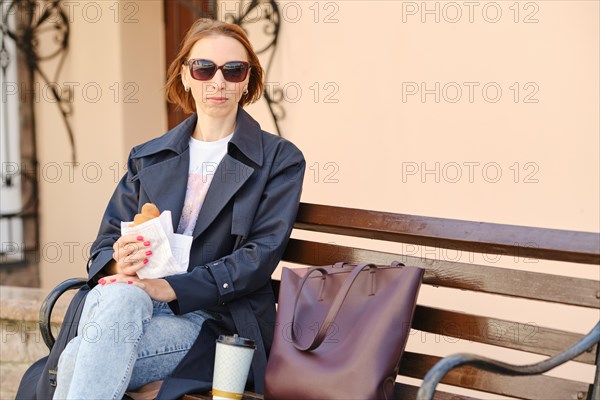Redhead woman enjoys cappuccino and croissant on city bench in stylish outfit