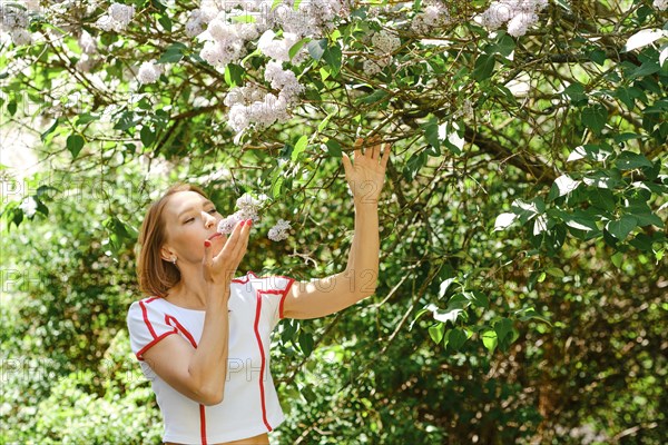 Adult redhead woman in tennis outfit sniffing lilac bush