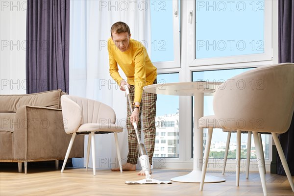 Middle-aged man washing floor with mop barefoot doing housework in modern apartment