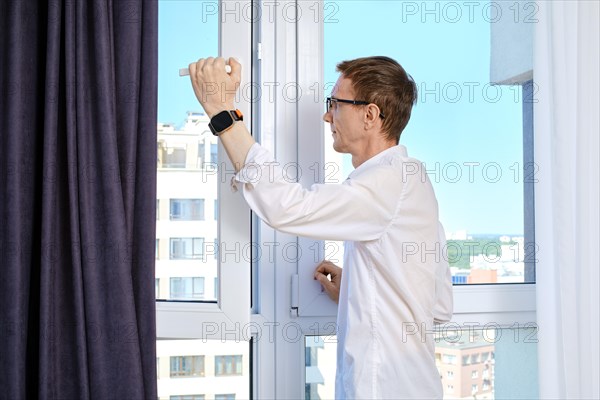 Middle aged man stands at open window of high-rise building