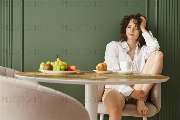 Dreamy woman in white shirt sitting at table in her modern apartment enjoying healthy fruit and morning coffee