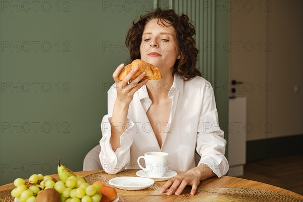 Young woman in shirt over her naked body sits at table in kitchen and looks at croissant she holds in her hand