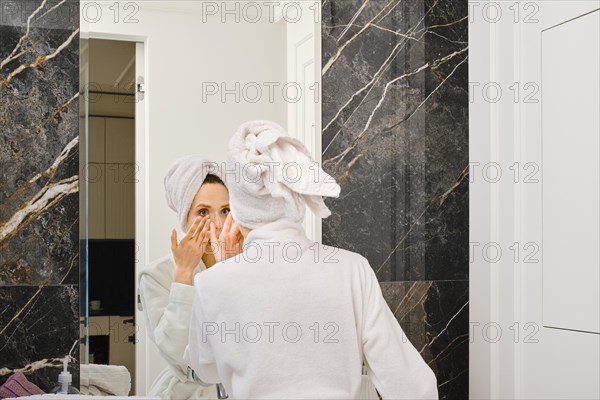 Young woman applying anti-wrinkle cream under her eyes after taking shower