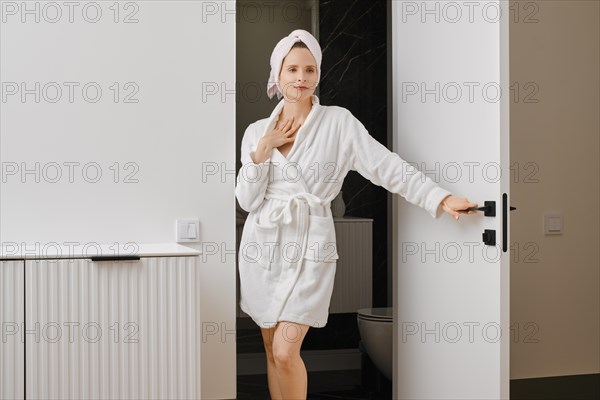 Pretty woman in a bathrobe and a towel on her head stands in the doorway of the bathroom