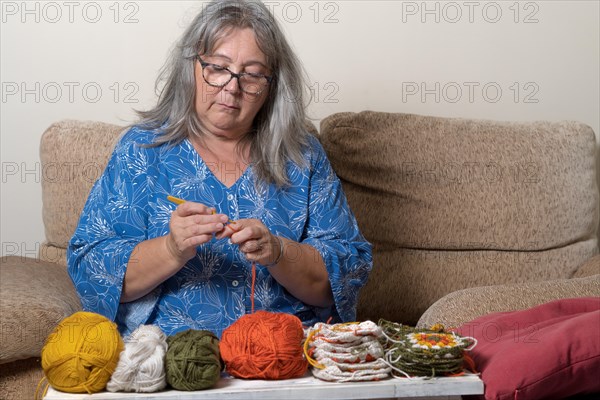 Front view of an older woman with white hair and glasses crocheting with her wool in the foreground