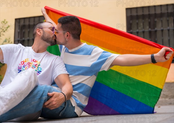 Portrait of gay male couple sitting on the floor kissing with rainbow flag at pride party