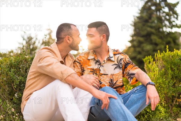 Romantic portrait of gay newlyweds sitting kissing at sunset in a park in the city