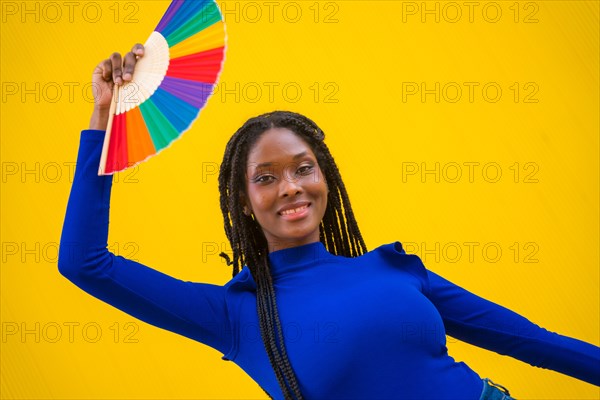 Portrait of a black ethnic woman dancing with a rainbow lgbt fan on a yellow background