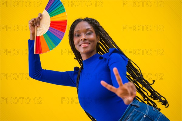 Portrait of a black ethnic woman dancing with a rainbow lgbt fan on a yellow background