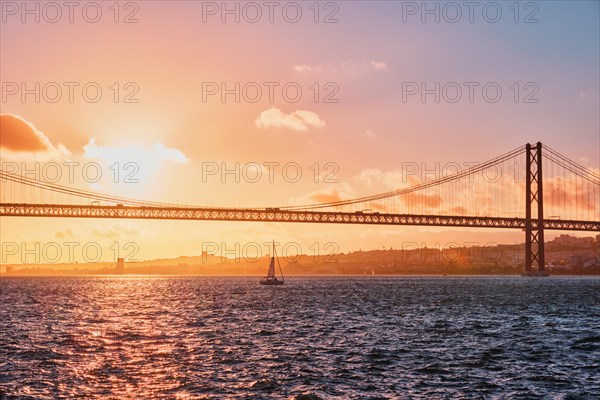 View of 25 de Abril Bridge famous tourist landmark of Lisbon connecting Lisboa and Almada on Setubal Peninsula over Tagus river with tourist yacht silhouette at sunset