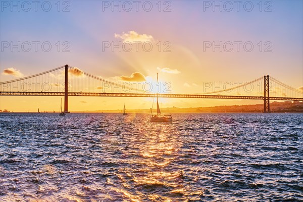 View of 25 de Abril Bridge famous tourist landmark of Lisbon connecting Lisboa and Almada on Setubal Peninsula over Tagus river with tourist yacht silhouette at sunset