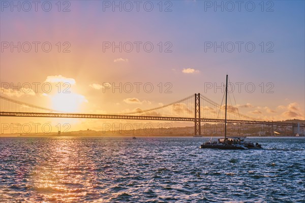 View of 25 de Abril Bridge famous tourist landmark of Lisbon connecting Lisboa and Almada on Setubal Peninsula over Tagus river with tourist yacht silhouette at sunset