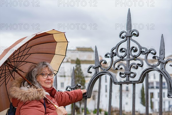 White-haired woman with coat and umbrella huddled against an iron fence on a dark and rainy autumn day