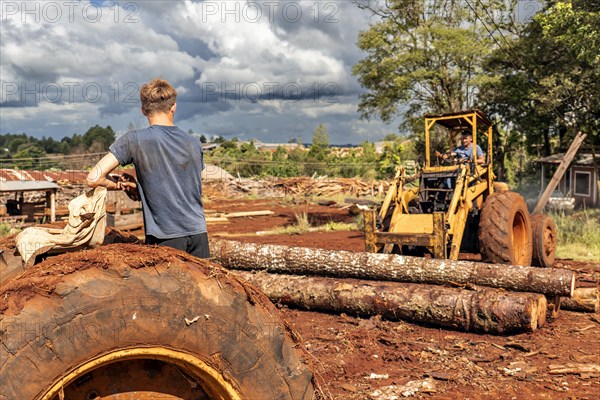 Man driving tractor at sawmill while his son watches him standing at another machine